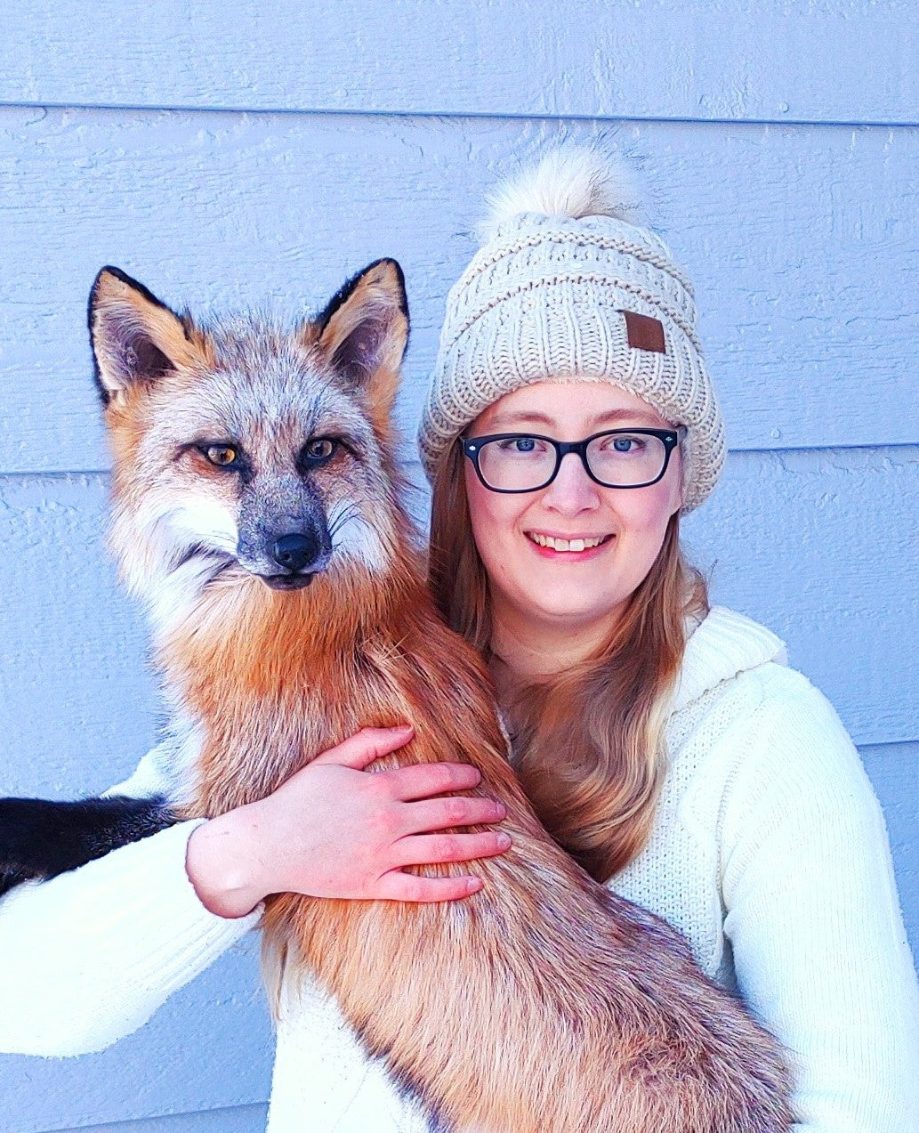 Person wearing a white beanie and glasses holding a fox against a light blue wall.